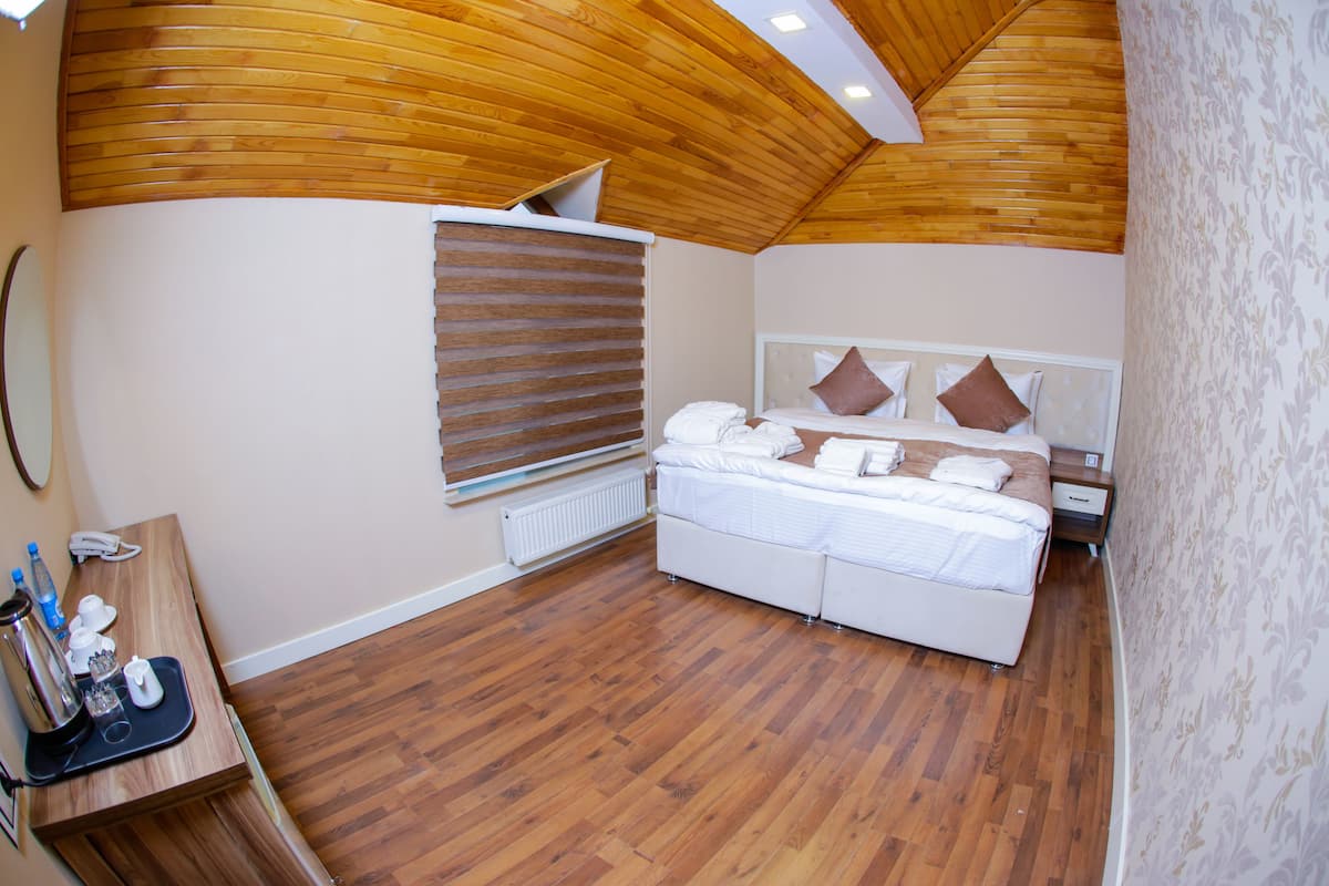 A bedroom with a wooden ceiling, a bed with white bedding, a bedside table with a lamp, and a window with blinds.