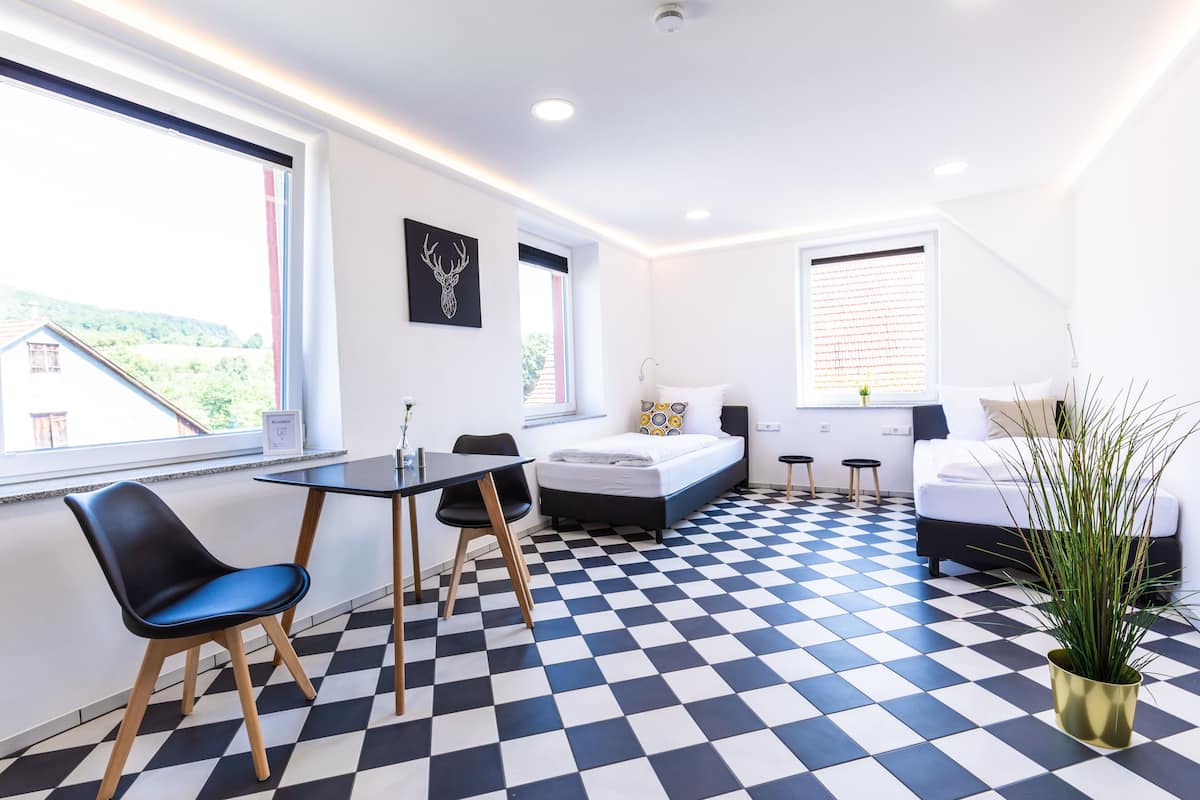 Une petite pièce avec un sol à carreaux, un lit, un bureau avec une chaise et une fenêtre donnant sur un paysage verdoyant.