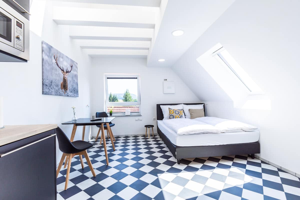 Une chambre moderne avec un sol à carreaux, un lit agrémenté d’oreillers à motifs, un bureau avec une chaise et un tableau représentant un cerf.