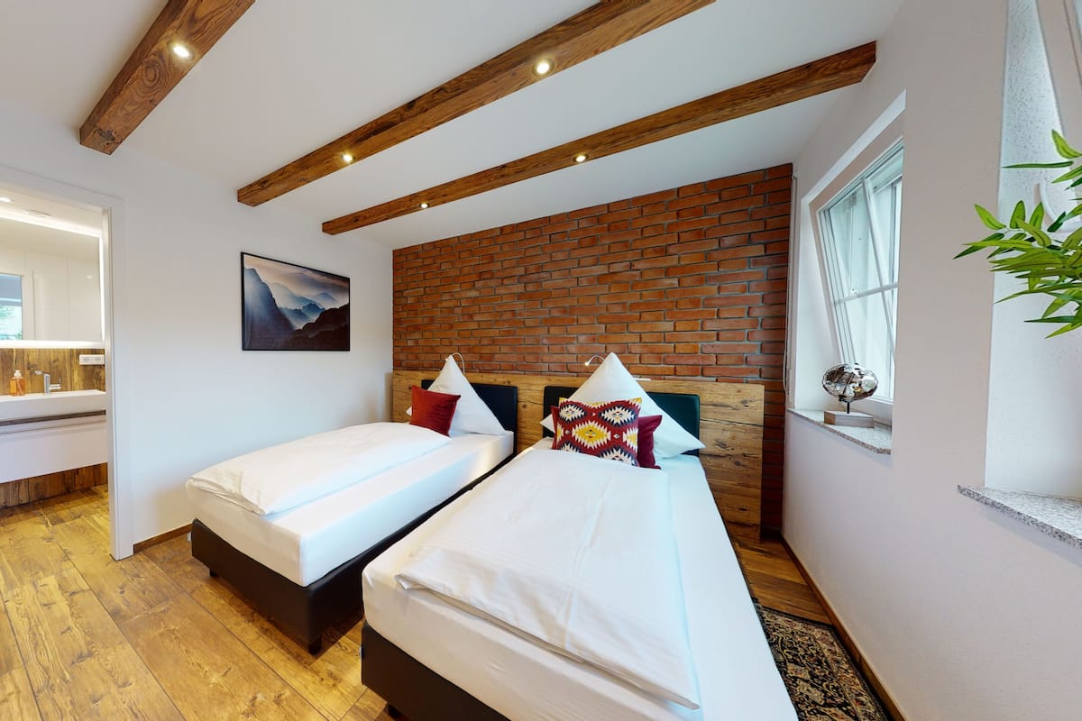Une chambre d’hôtel moderne avec deux lits, un plafond en bois, un mur en briques et une salle de bain équipée d’un miroir et d’un lavabo.