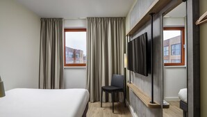 Habitación de hotel con cama, una silla, televisor montado en la pared y ventana con cortinas.