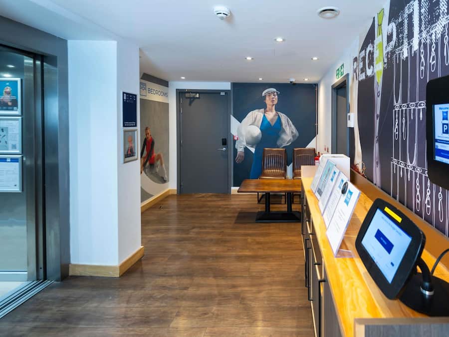 Un vestíbulo de hotel con mostrador de recepción, un cartel grande de una mujer con vestido azul y una señal que indica el camino a las habitaciones.