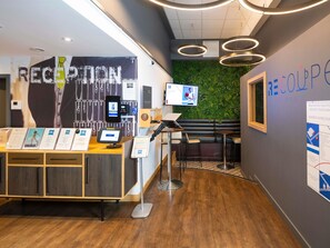 A reception area with a desk, seating, a green wall, and a TV screen. - ibis budget Swindon Town Centre (Swindon)
