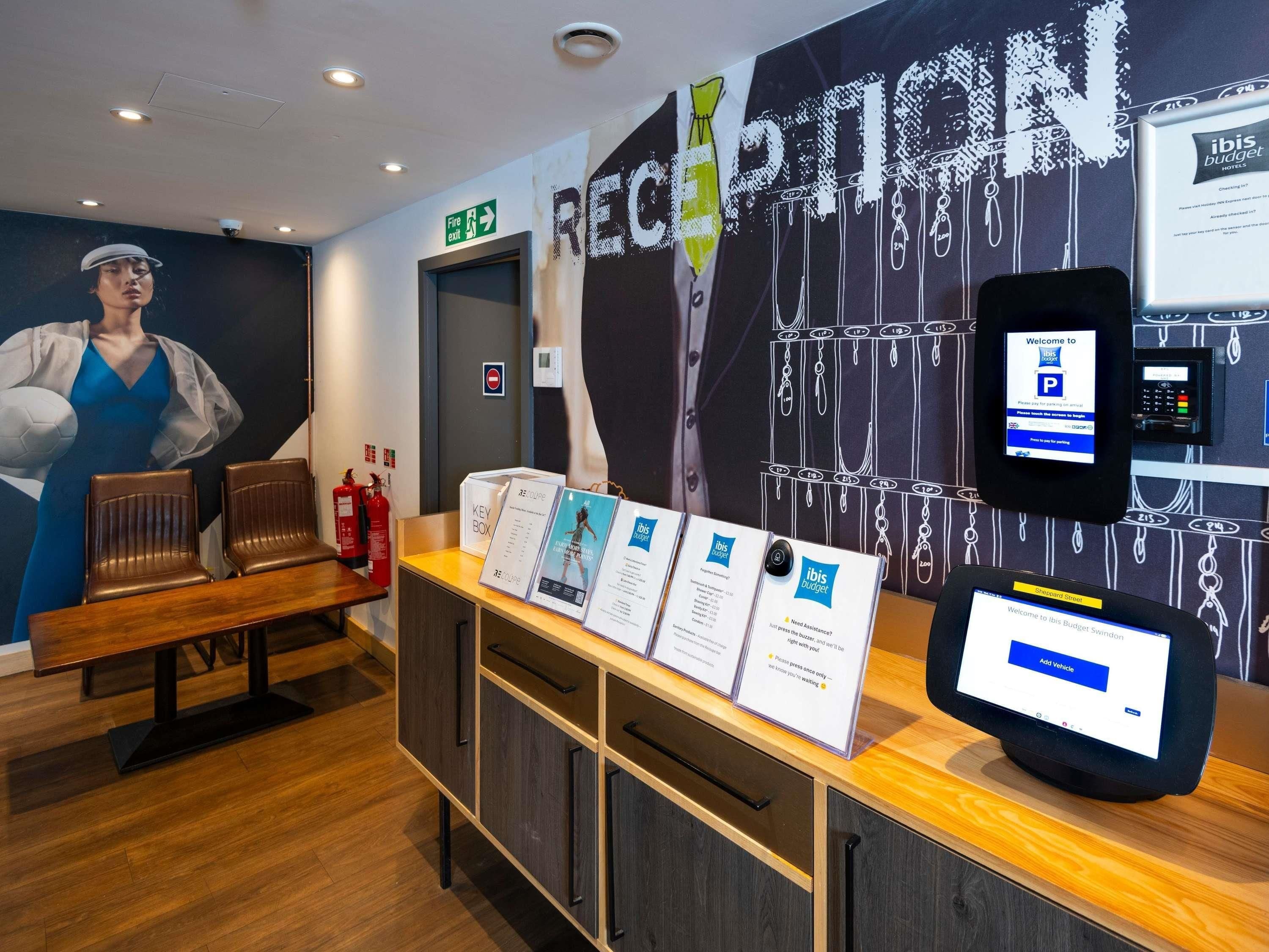A reception area with a desk, chairs, a fire exit sign, a key box, and a digital display for hotel services.