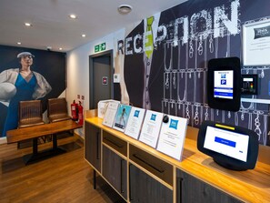 Zona de recepción con mostrador, sillas, señal de salida de incendios, caja para llaves y pantalla digital con servicios del hotel.