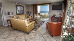 A living room with a beige sofa, a coffee table, and a TV. There is a balcony with a view of the ocean. - 110 South Hampton - Barefoot Resort Rentals (Myrtle Beach)