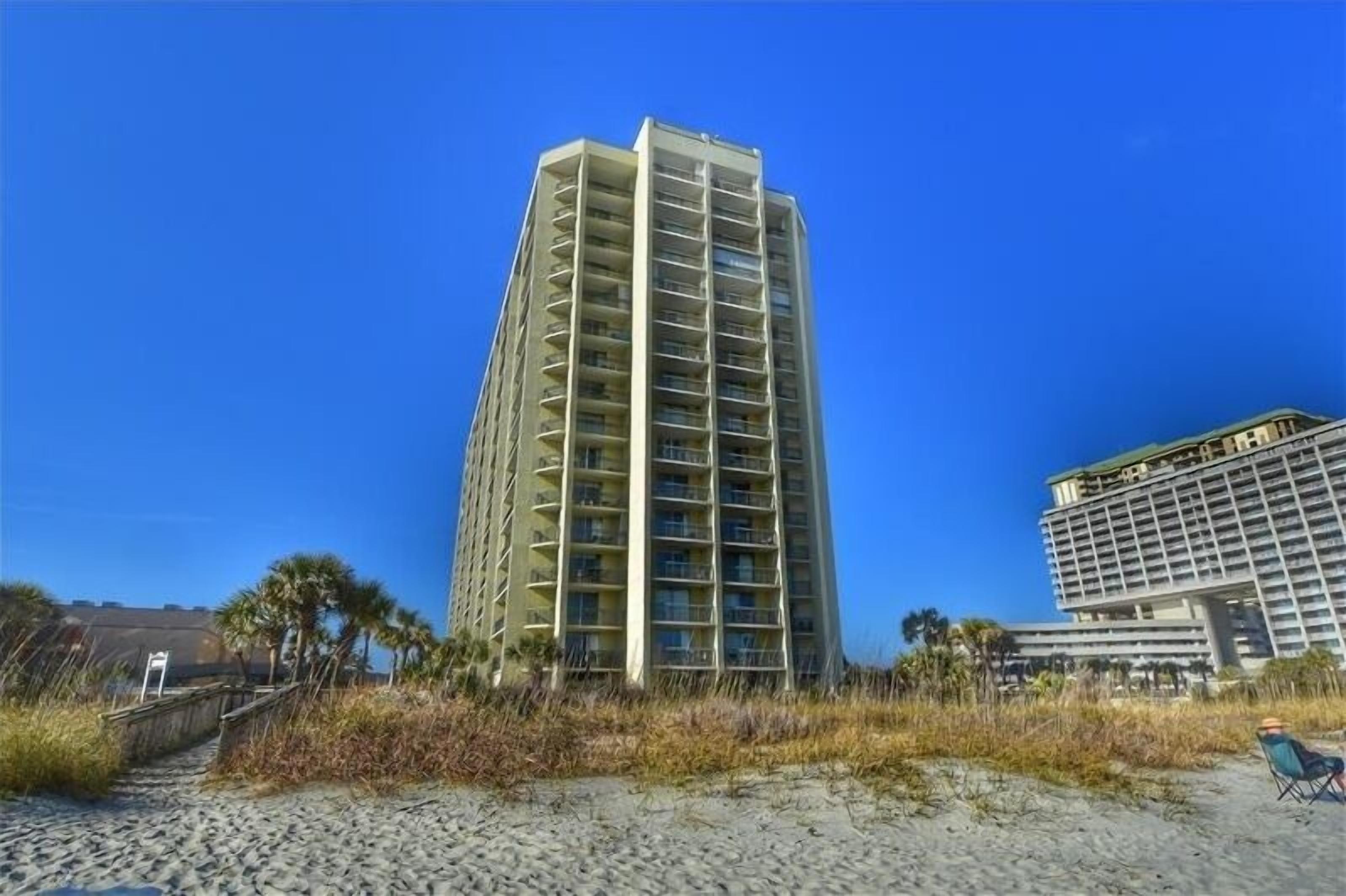 A tall hotel with multiple floors and balconies, located near a beach with a sandy path leading to the entrance.