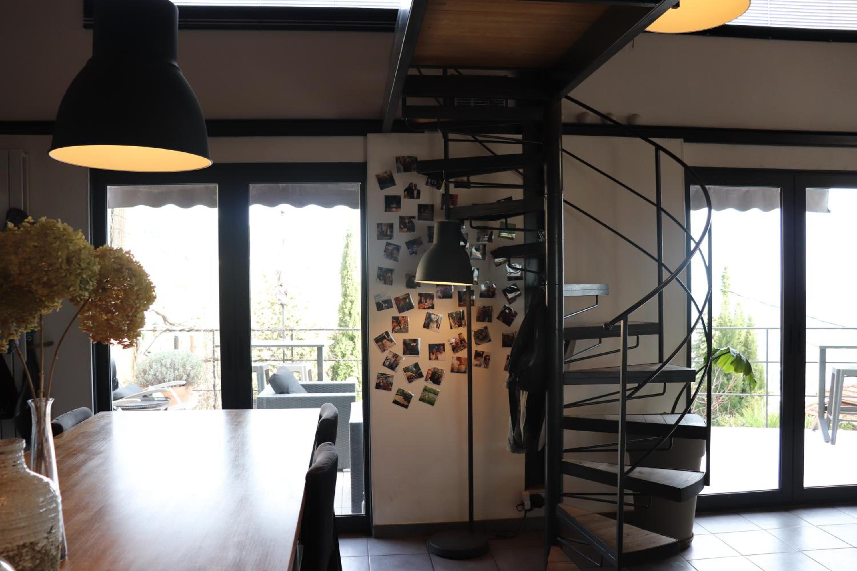 Un interior moderno con una mesa de comedor de madera, un jarrón con flores secas, una escalera y una pared decorada con fotografías.