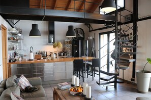 A modern kitchen with a central island, a dining area with chairs, a staircase, and a living room with a sofa and a coffee table. - Skylark Bed & Breakfast (Grasse)