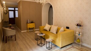 Una sala de estar con un sofá amarillo, dos mesitas auxiliares, una araña y una estantería decorativa.