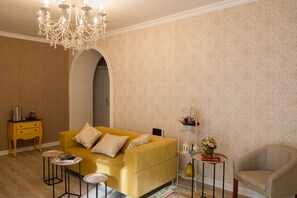 Un salon avec un canapé jaune, un lustre, une petite table d’appoint et un fauteuil à motifs.
