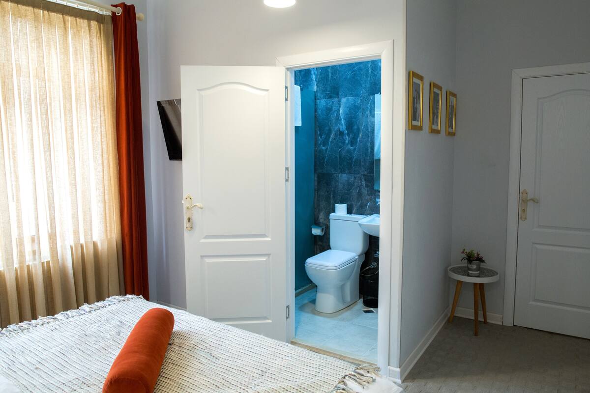 Un dormitori amb un llit, una finestra amb cortines, un bany amb vàter i rentamans, i una porta que condueix a una altra habitació.