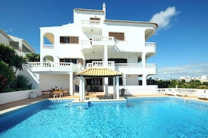 Un edificio blanco de varios pisos con piscina, zona de estar exterior y un patio cubierto.
