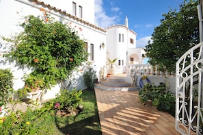 Um pátio com um edifício branco, um portão branco e um pequeno jardim com diversas plantas.
