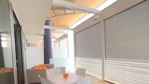 A modern outdoor dining area with white chairs and a table, a blue pillar, and a closed roller shutter door. - Bilocale Marina (Gallipoli)