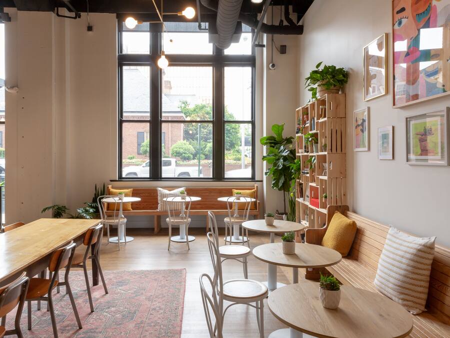 Una cafetería con una larga mesa de madera, varias sillas, un banco y una estantería con plantas.