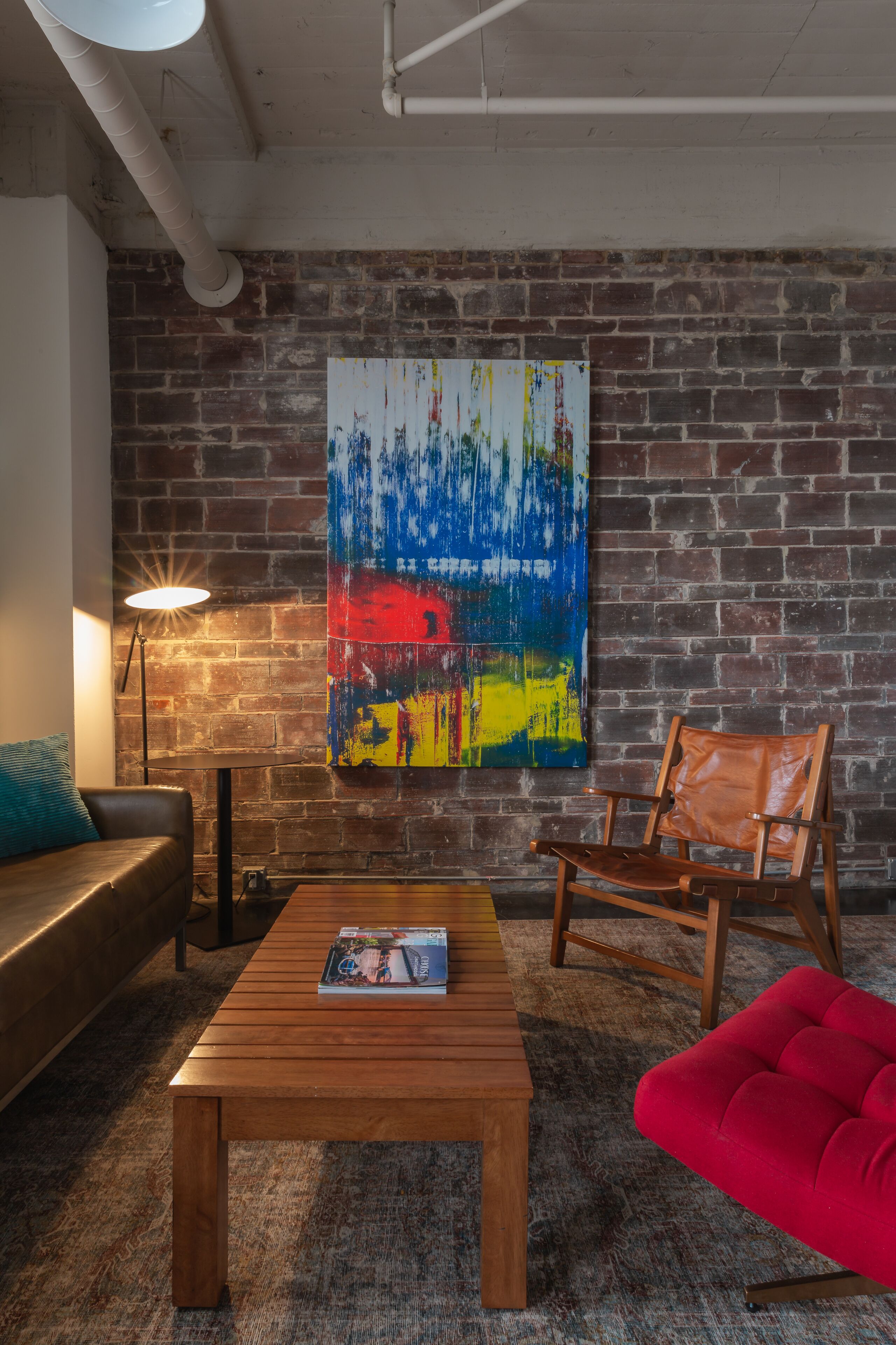 A room with a brick wall, a colorful abstract painting, a wooden coffee table, a red sofa, and a leather couch.