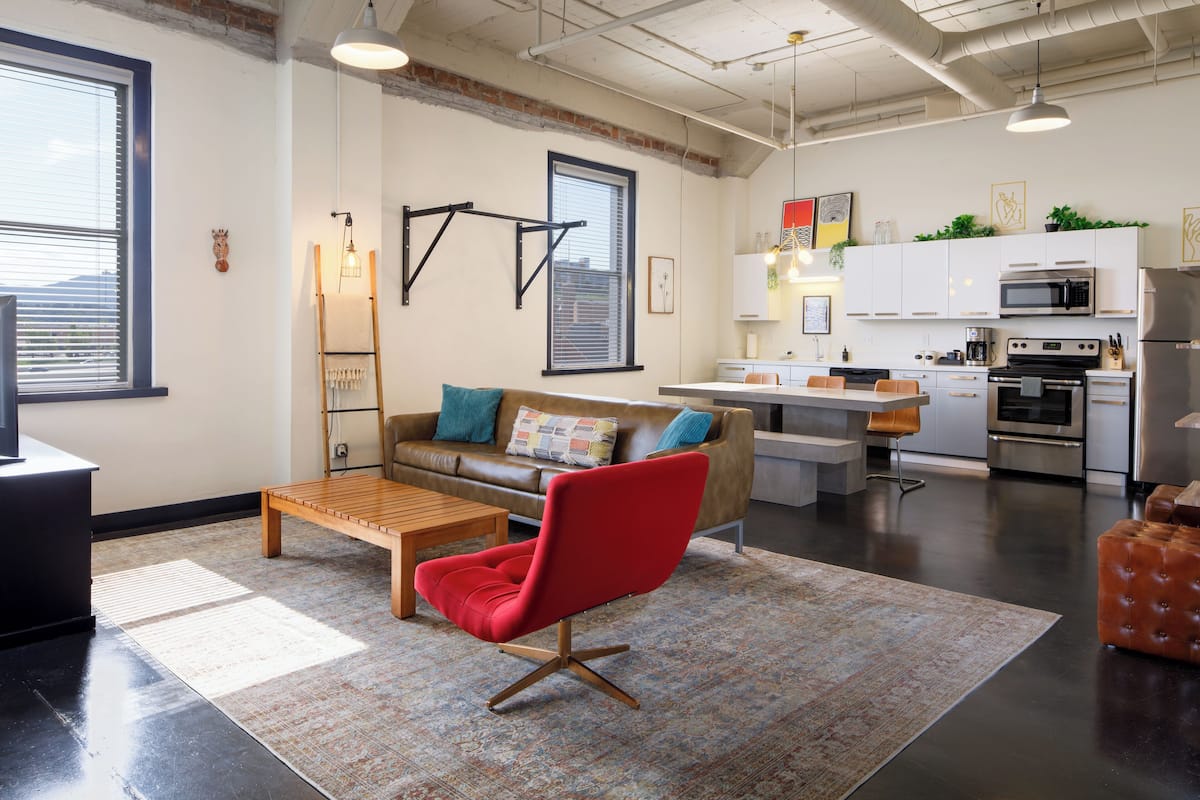 A modern living room with a red chair, a wooden coffee table, and a sofa with colorful cushions. There is a kitchen area with white cabinets and stainless steel appliances.