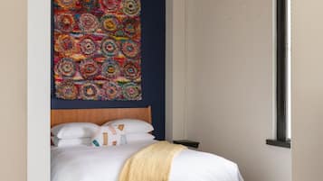 A bedroom with a wooden bed, white bedding, a colorful abstract wall hanging, and a hanging light fixture.