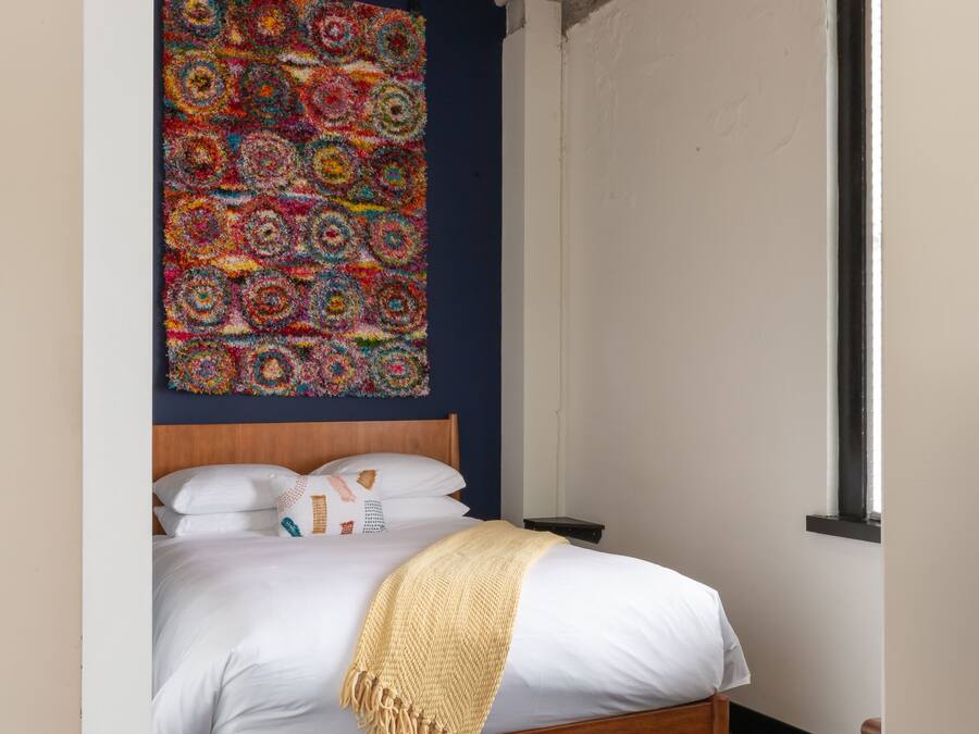 A bedroom with a wooden bed, white bedding, a colorful abstract wall hanging, and a hanging light fixture.