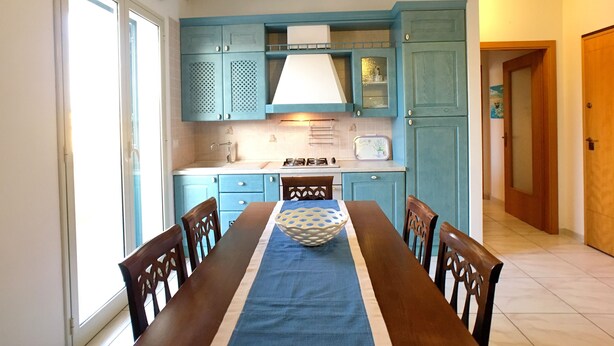 A dining area with a table set for four, a kitchen with teal cabinets, and a window providing natural light. - Bilocale Rosalba (Gallipoli)