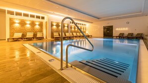 An indoor swimming pool with a metal ladder, lounge chairs, and a stone wall with lights.