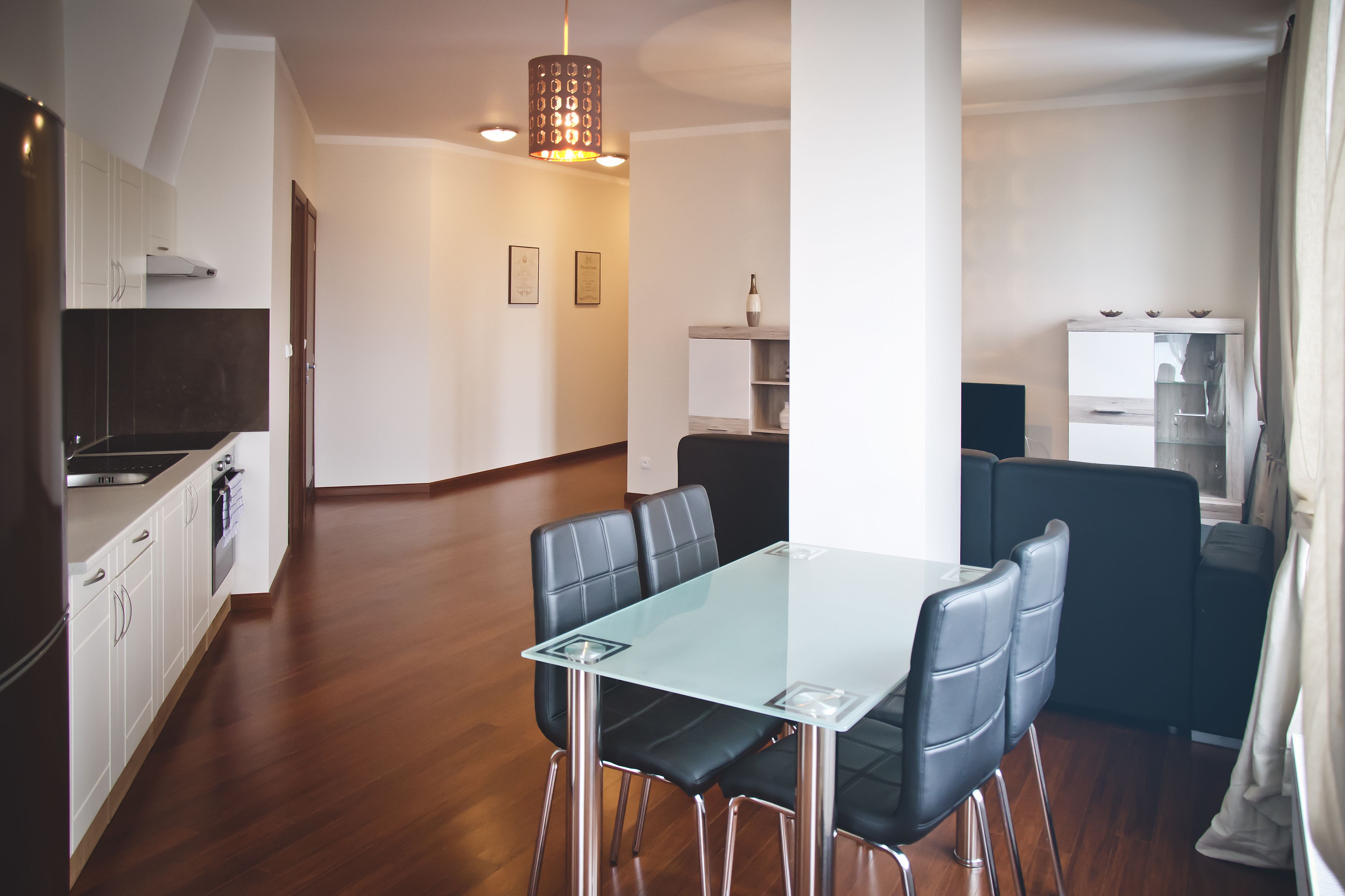A modern kitchen with white cabinets, a black countertop, and stainless steel appliances. There is a dining area with a glass table and black chairs.