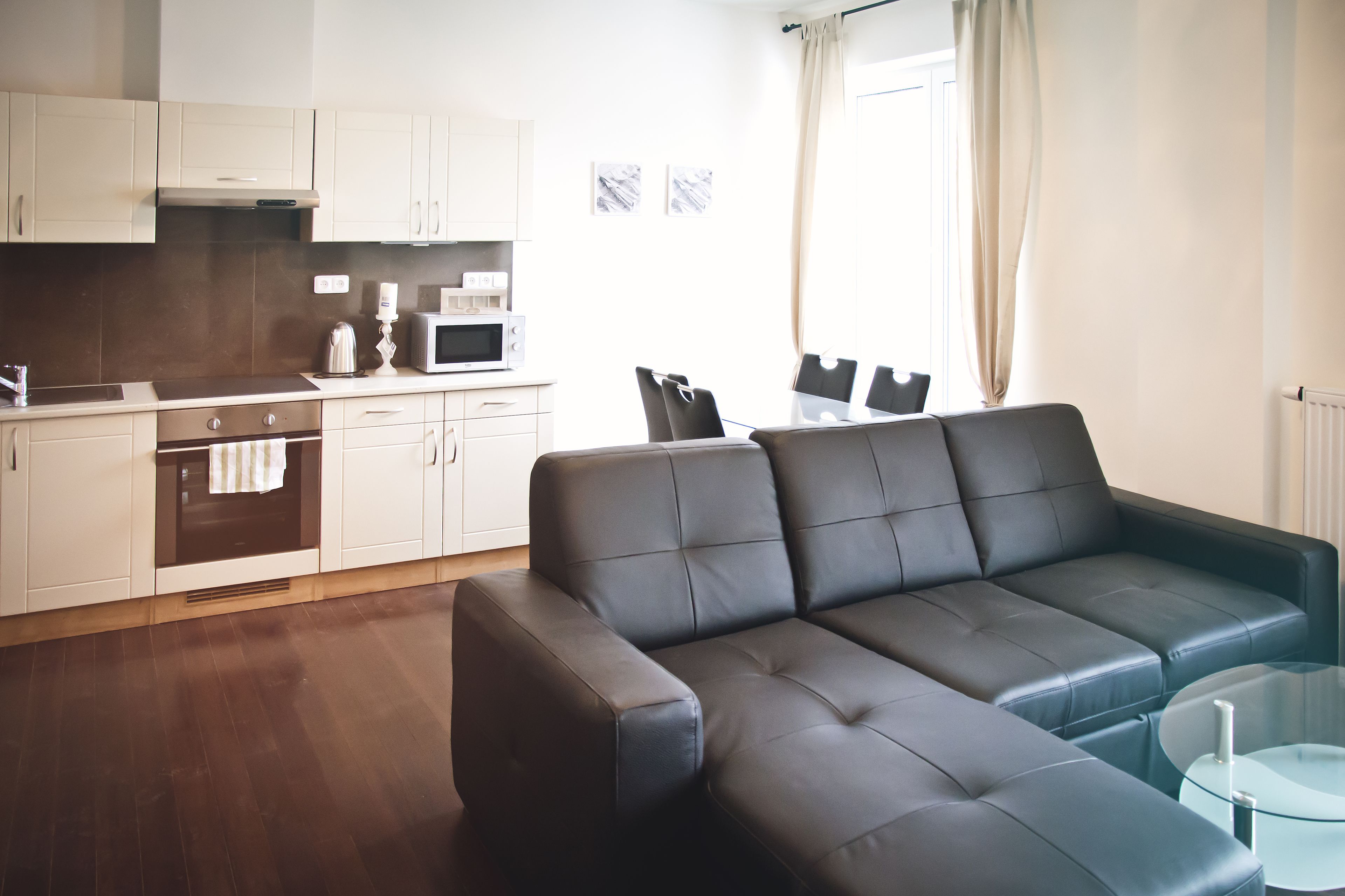 Ein modernes Wohnzimmer mit einer schwarzen Ledersofa, einem Glastisch und einer Küchenzeile mit weißen Schränken und einer Mikrowelle.