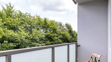 Een balkon met een houten stoel, een kleine tafel met een fles en fruit, en uitzicht op groene bomen.