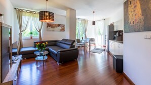 A modern living room with a sectional sofa, a glass coffee table, and a dining area with a small table and chairs. There is a kitchen area with cabinets and a microwave.
