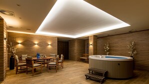 A modern indoor space with a wooden floor, a round dining table with chairs, a hot tub, and wall-mounted lighting.
