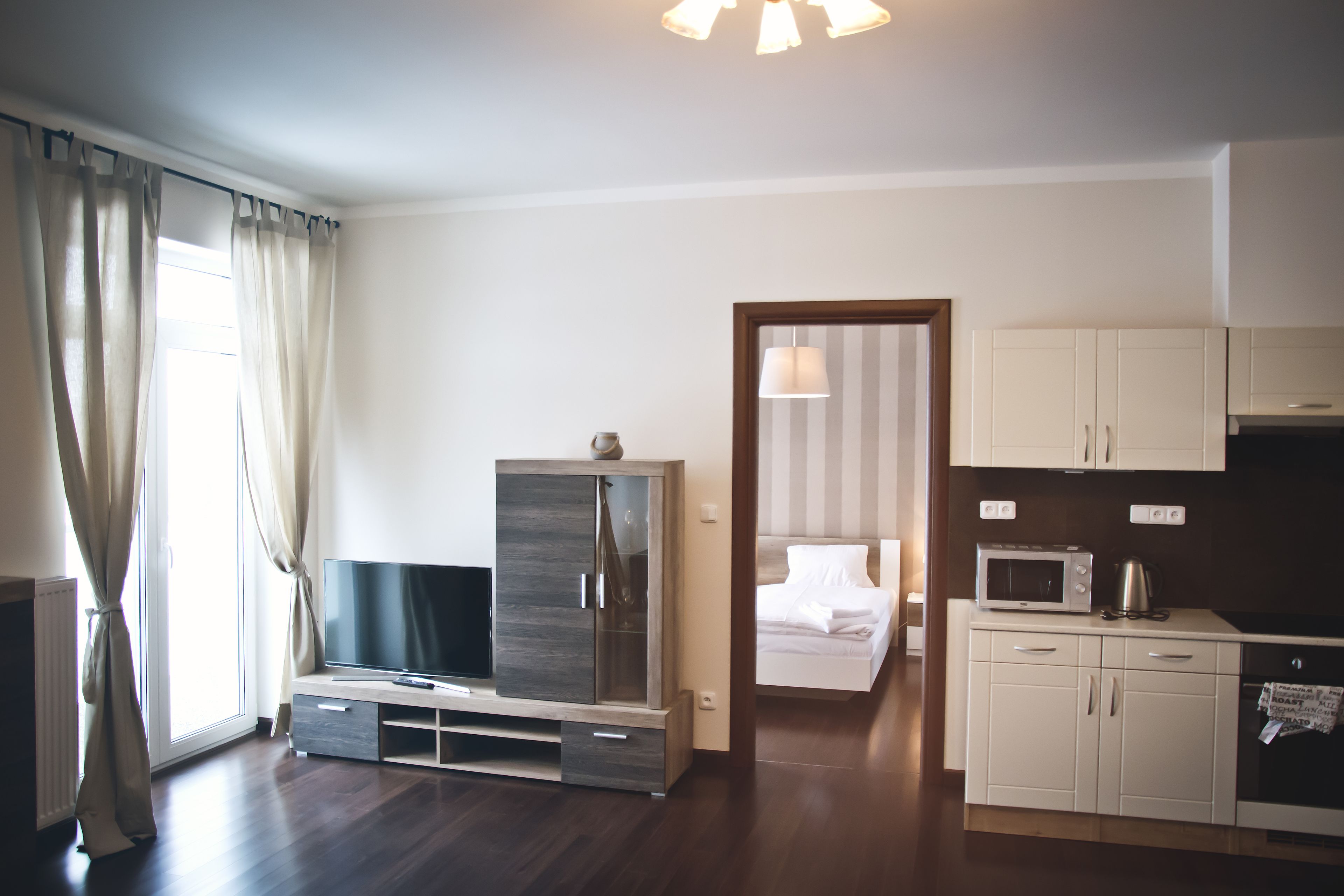 A modern hotel room with a flat-screen TV, a wardrobe, a bed, a kitchenette, and a large window with curtains.