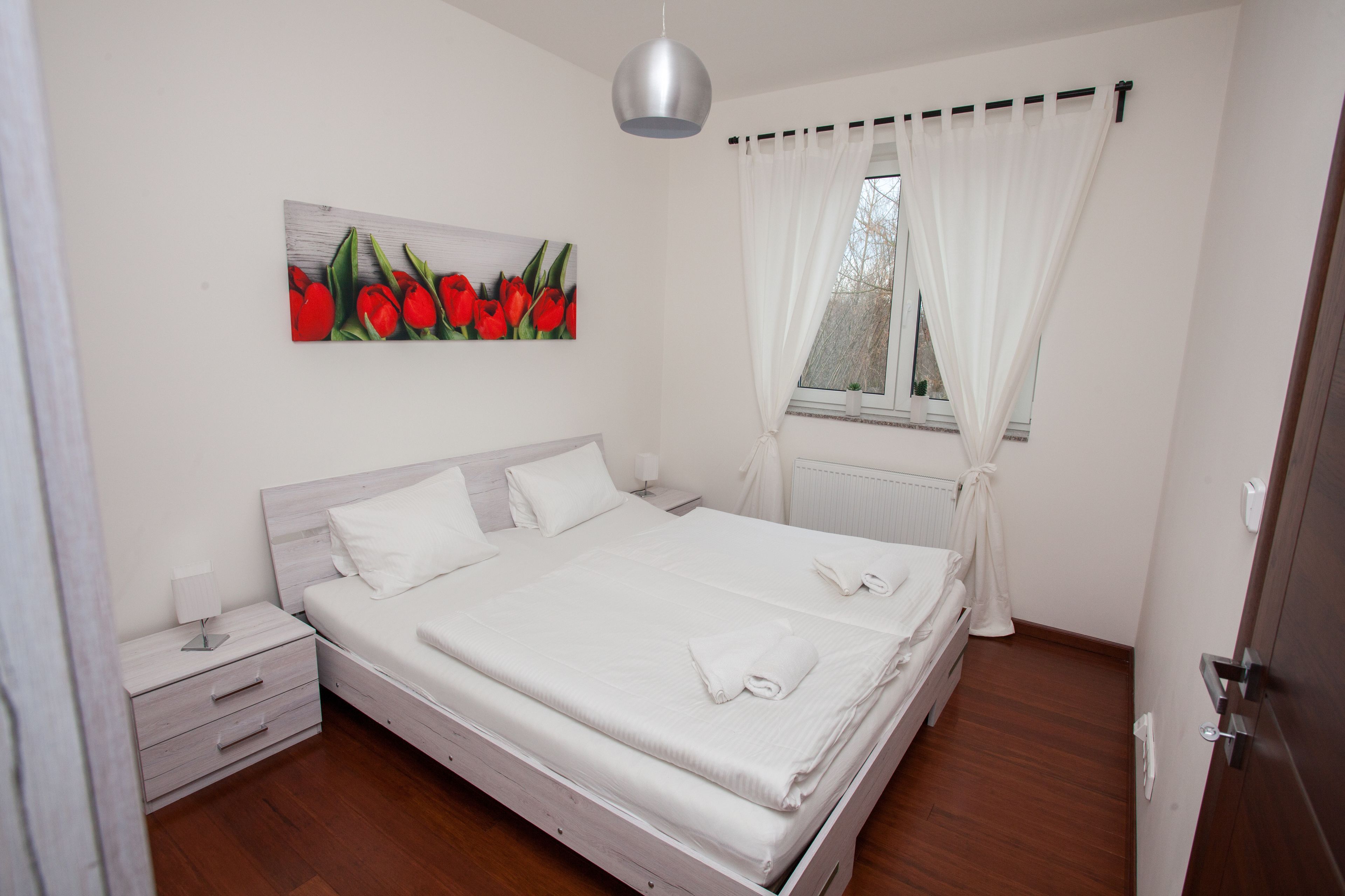 Una cama bien tendida con sábanas blancas y dos almohadas, una mesita de noche con una lámpara, un cuadro colgado en la pared de tulipanes rojos, y una ventana con cortinas blancas.