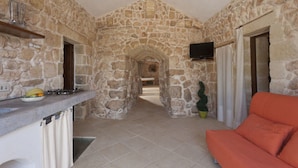 A stone-walled interior with a kitchen area, a TV mounted on the wall, and a seating area with a sofa. - Bilocale Rurale in Resort (Morciano di Leuca)