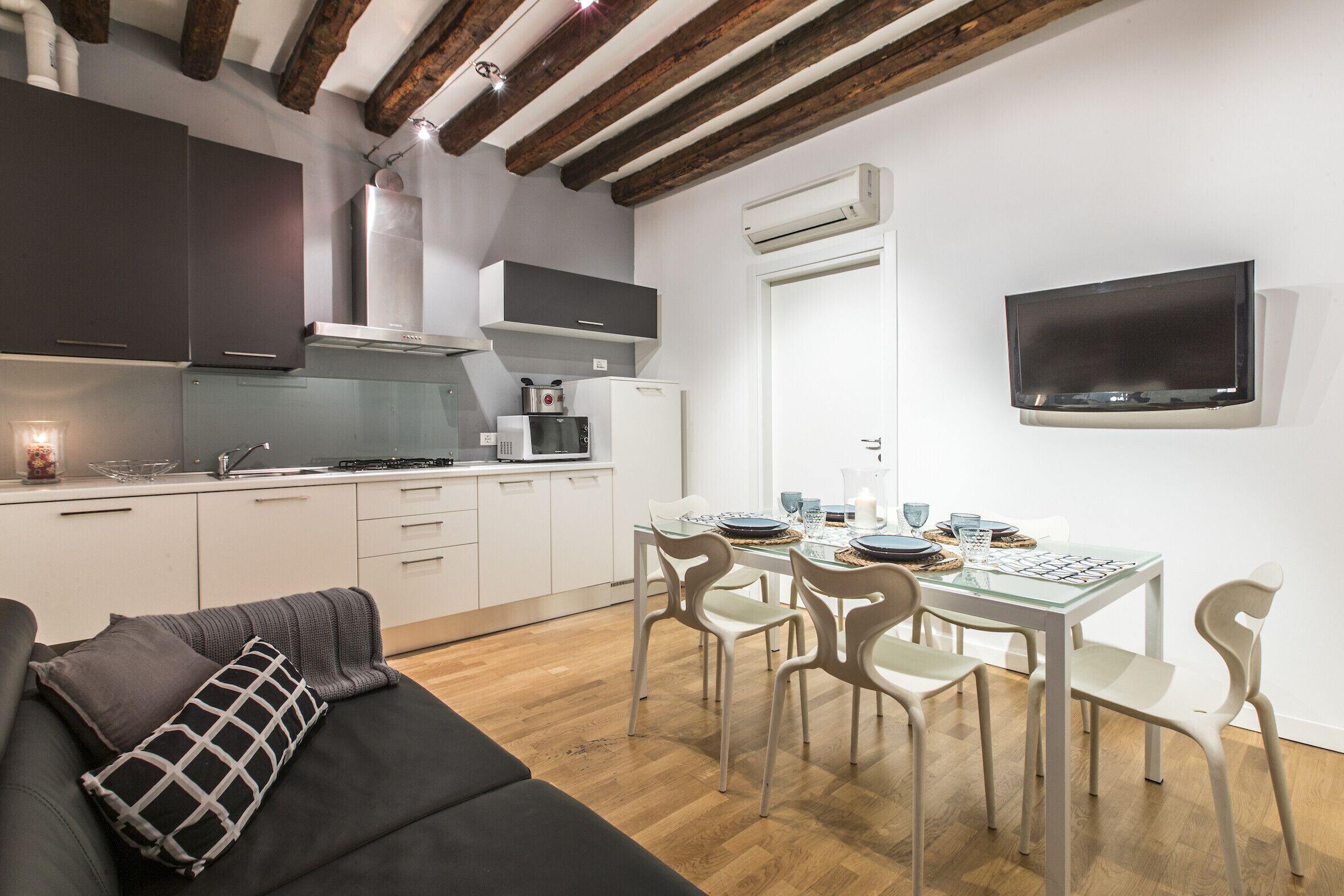 A modern kitchen with white cabinets, a dining area with a glass table and white chairs, and a flat-screen TV mounted on the wall.