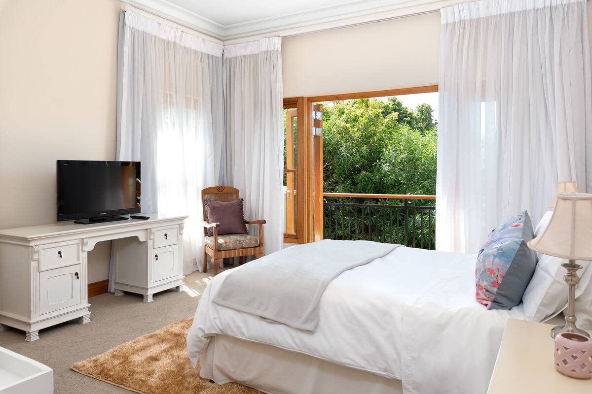 Un dormitorio con cama, escritorio con televisor, silla, balcón con vistas a zonas verdes y una lámpara.