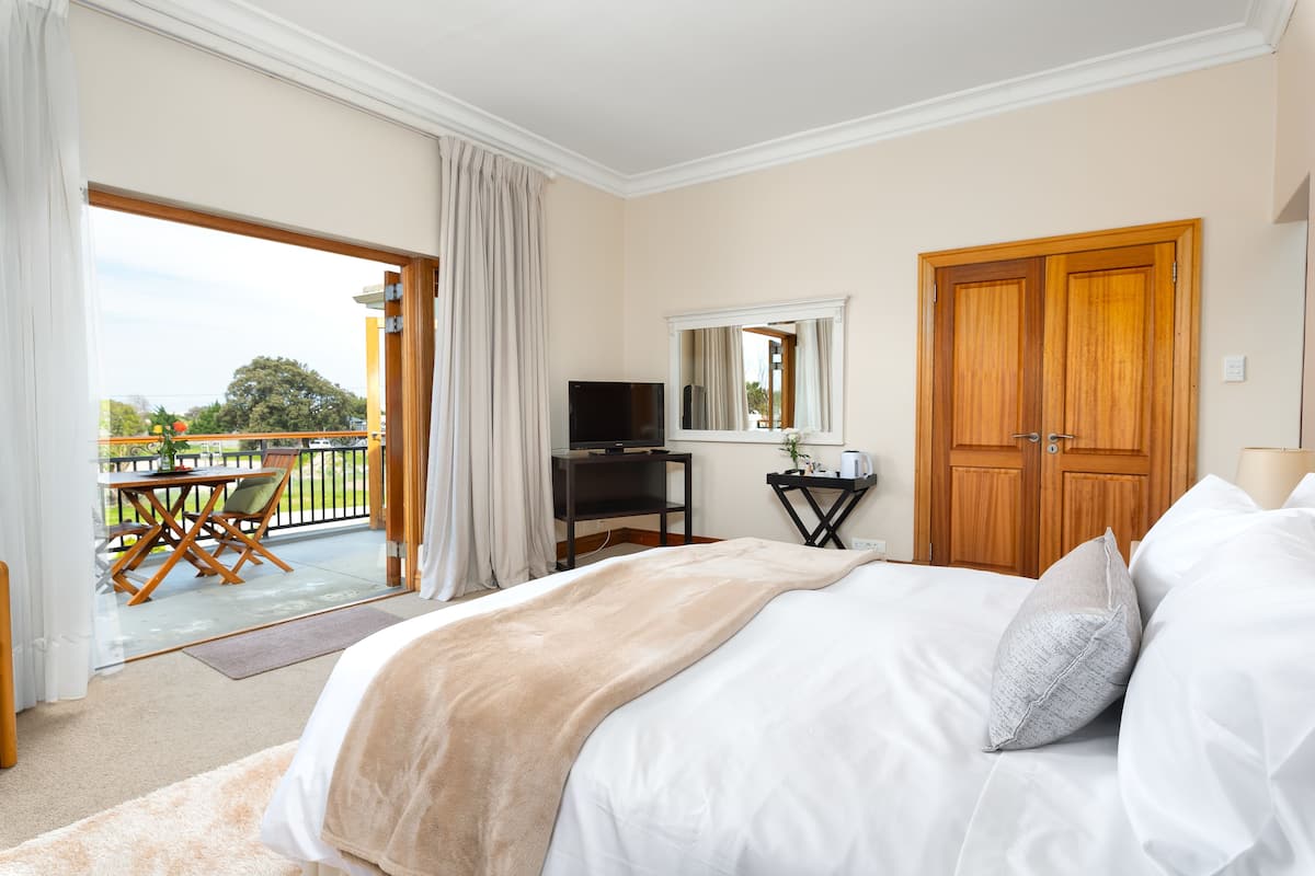 Un dormitorio con una cama grande, televisión, armario de madera, balcón con mesa y sillas, y vista al exterior.
