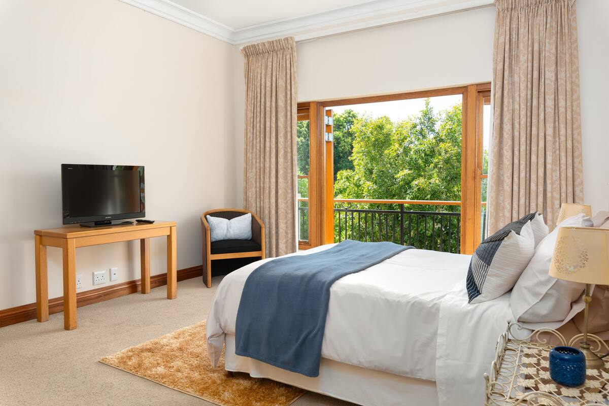 Un dormitorio con cama, televisión sobre una mesa de madera, una silla y un balcón con vistas a árboles.