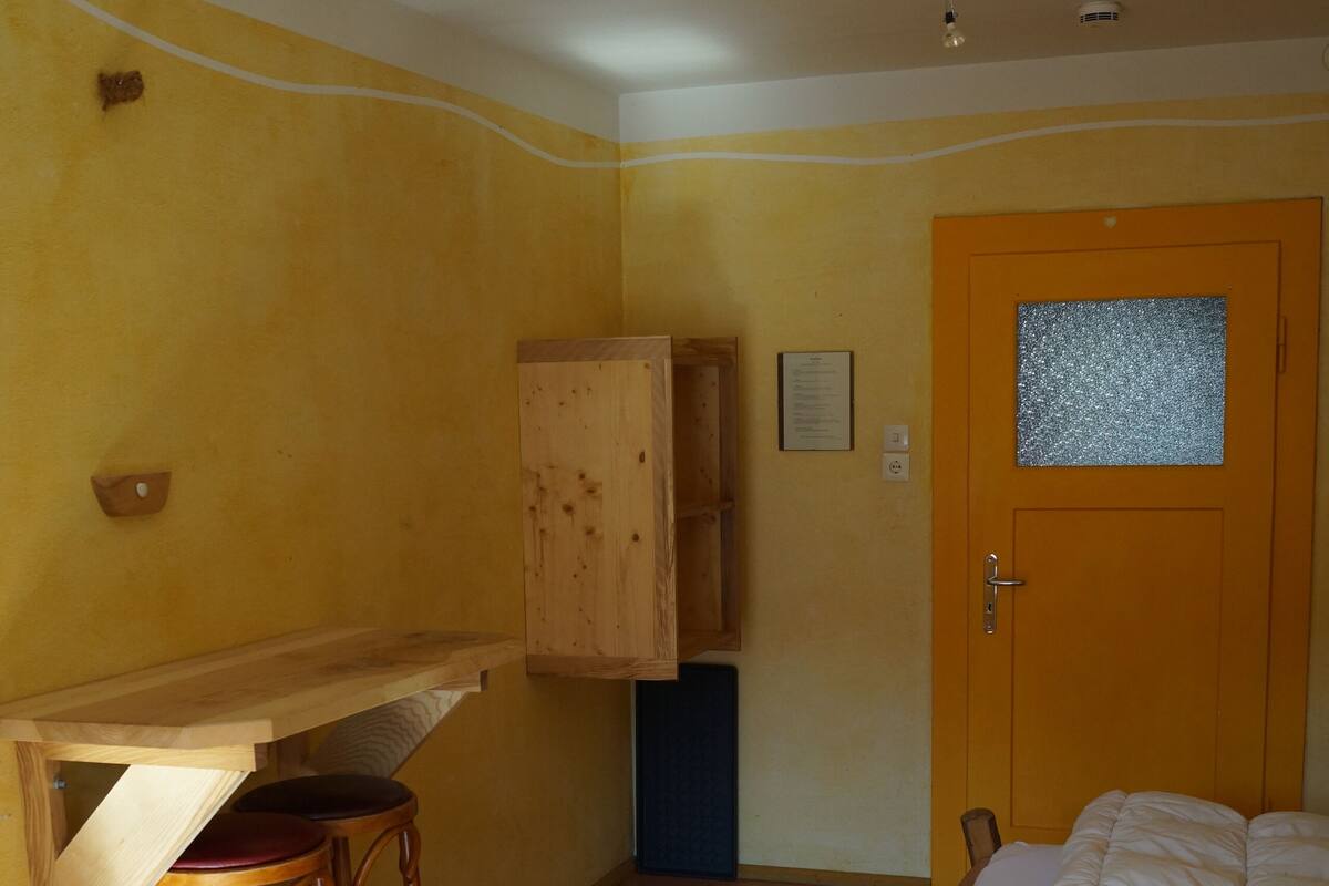 Una habitación con un escritorio de madera, dos taburetes altos, una cama con ropa blanca, una puerta de madera con ventanal y una lámpara en el techo.
