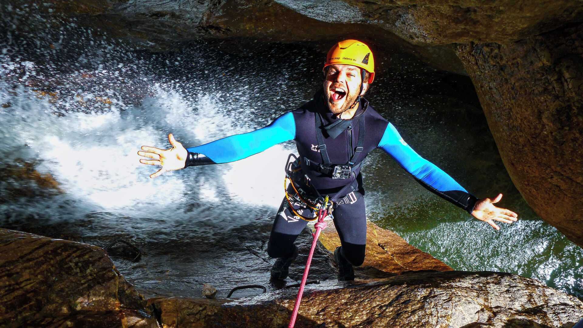 A person in a cave with a helmet and harness, standing on a rock with water splashing around.