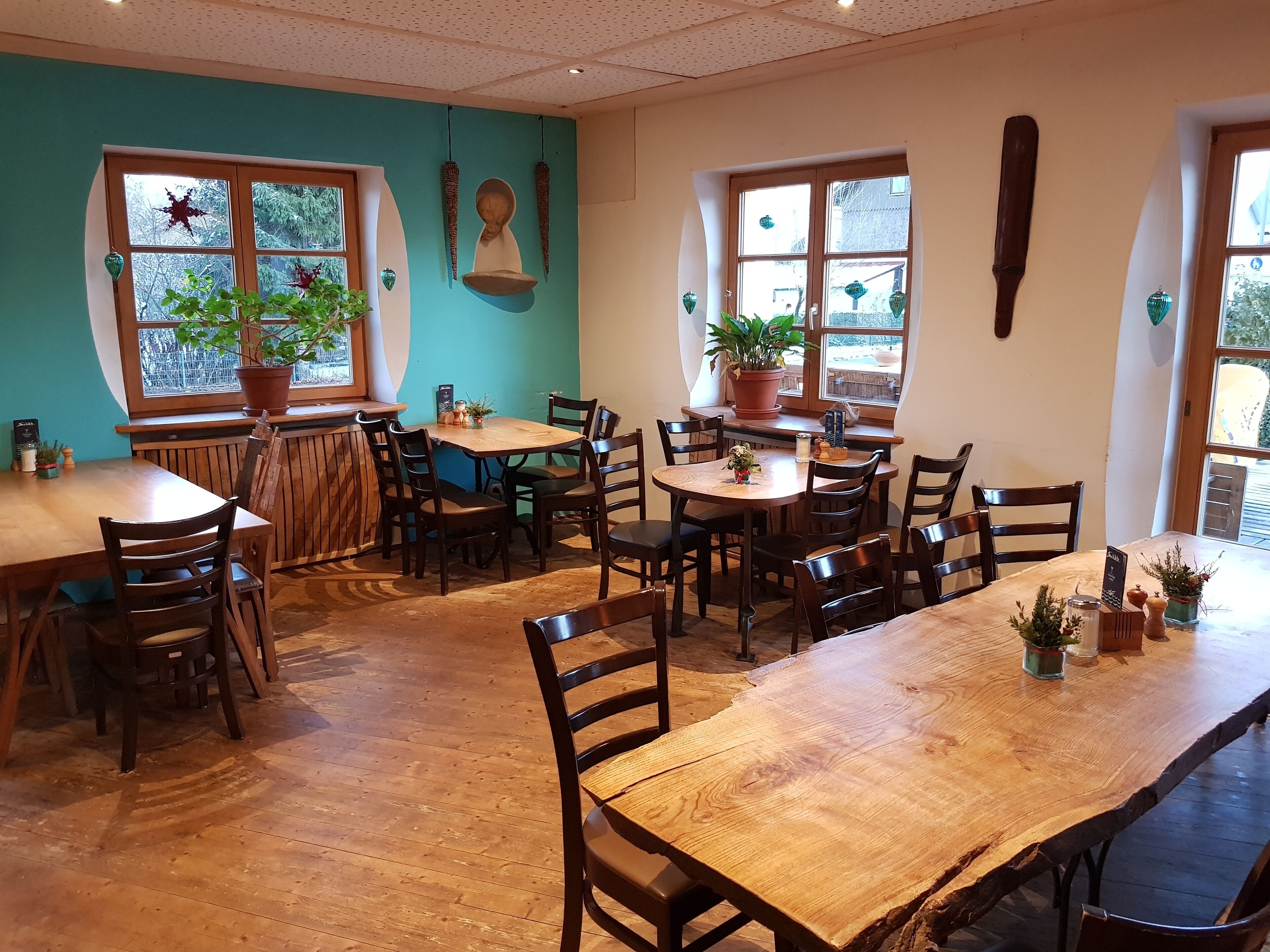A cozy restaurant with wooden tables and chairs, potted plants, and large windows.