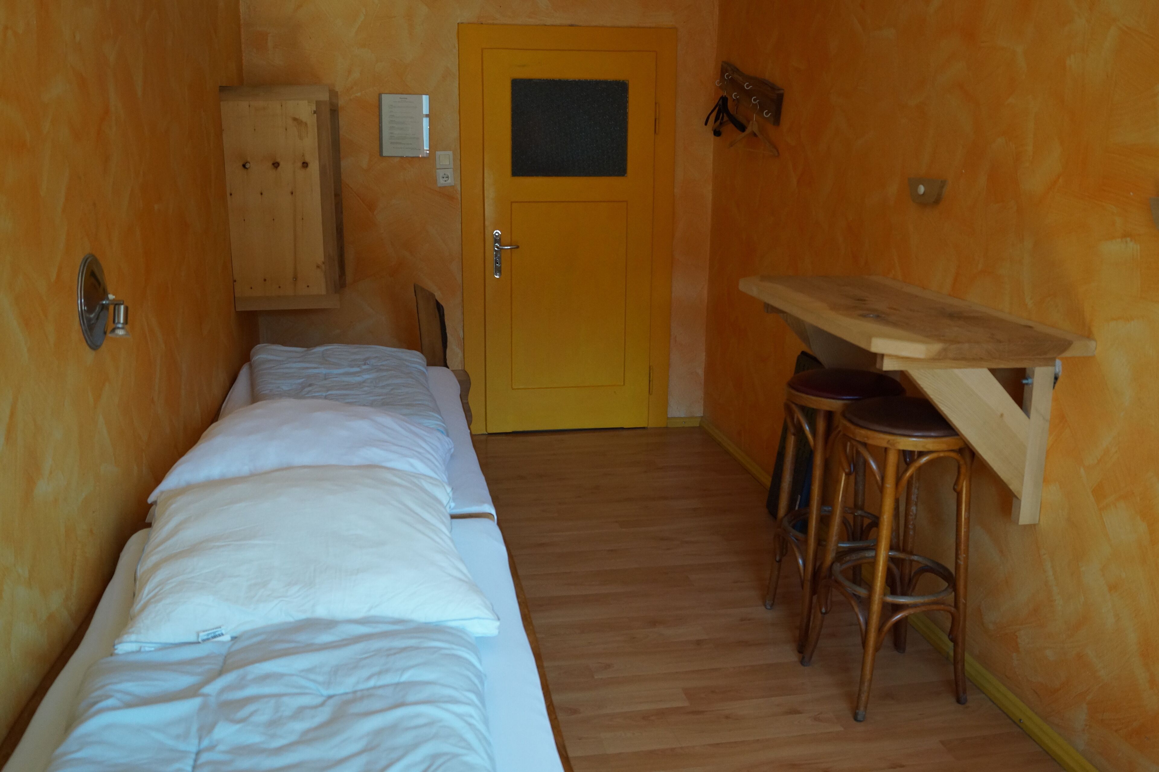 Ein schmaler Raum mit einem hölzernen Bett, einem Schrank aus Holz und einem kleinen Holztisch mit Hockern.