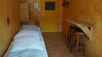 Ein schmaler Raum mit einem hölzernen Bett, einem Schrank aus Holz und einem kleinen Holztisch mit Hockern.