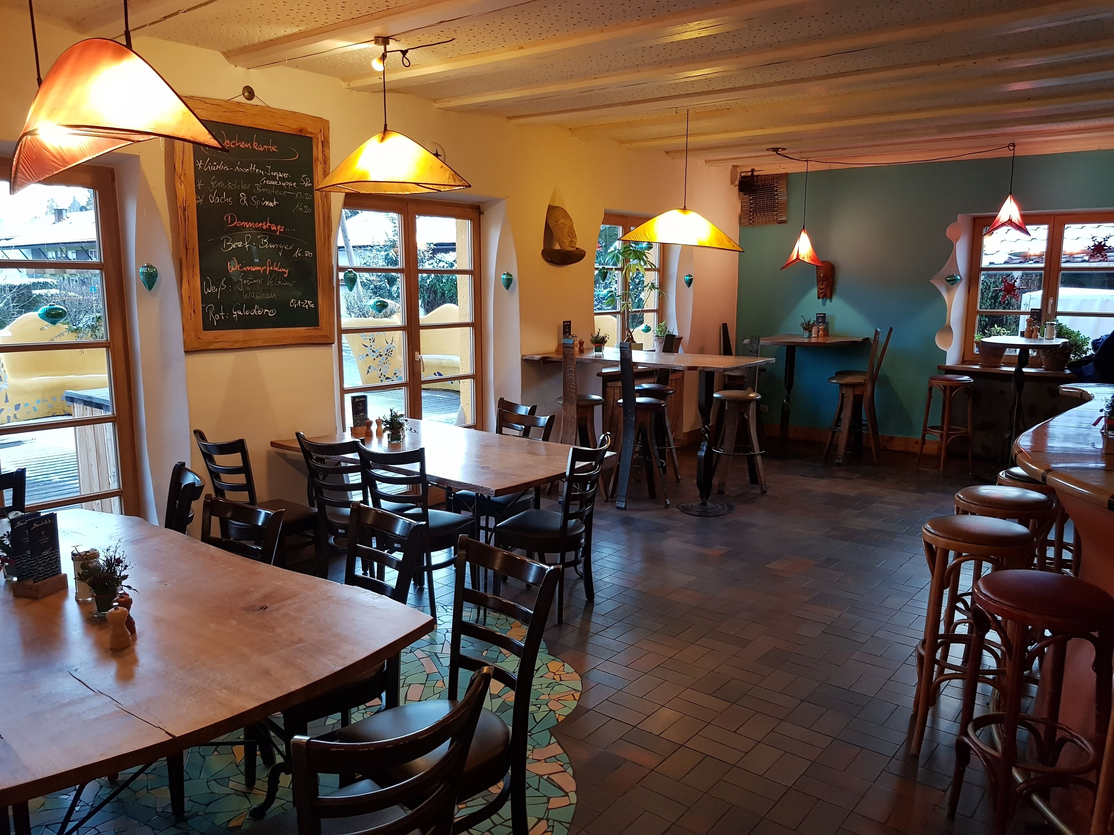 Ein Restaurantinnenraum mit Holztischen und -stühlen, hängenden Pendelleuchten und einer Kreidetafel mit Speisekarte.