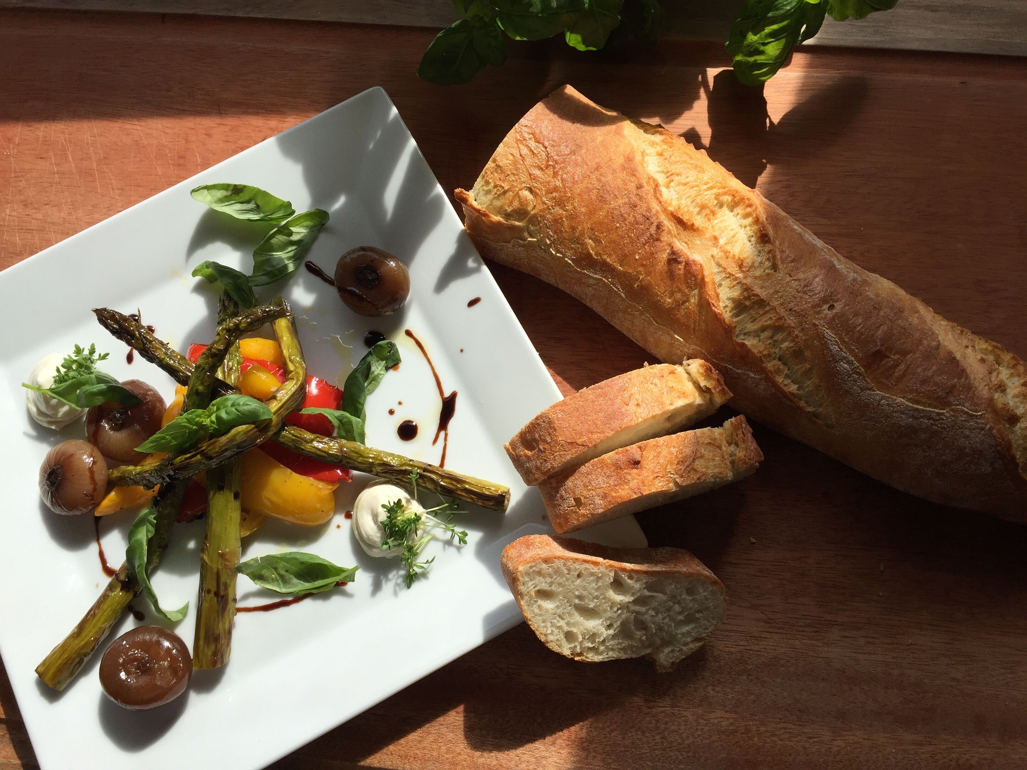 A white plate with a vegetable dish, including asparagus, tomatoes, and a small dollop of cream, accompanied by a baguette.