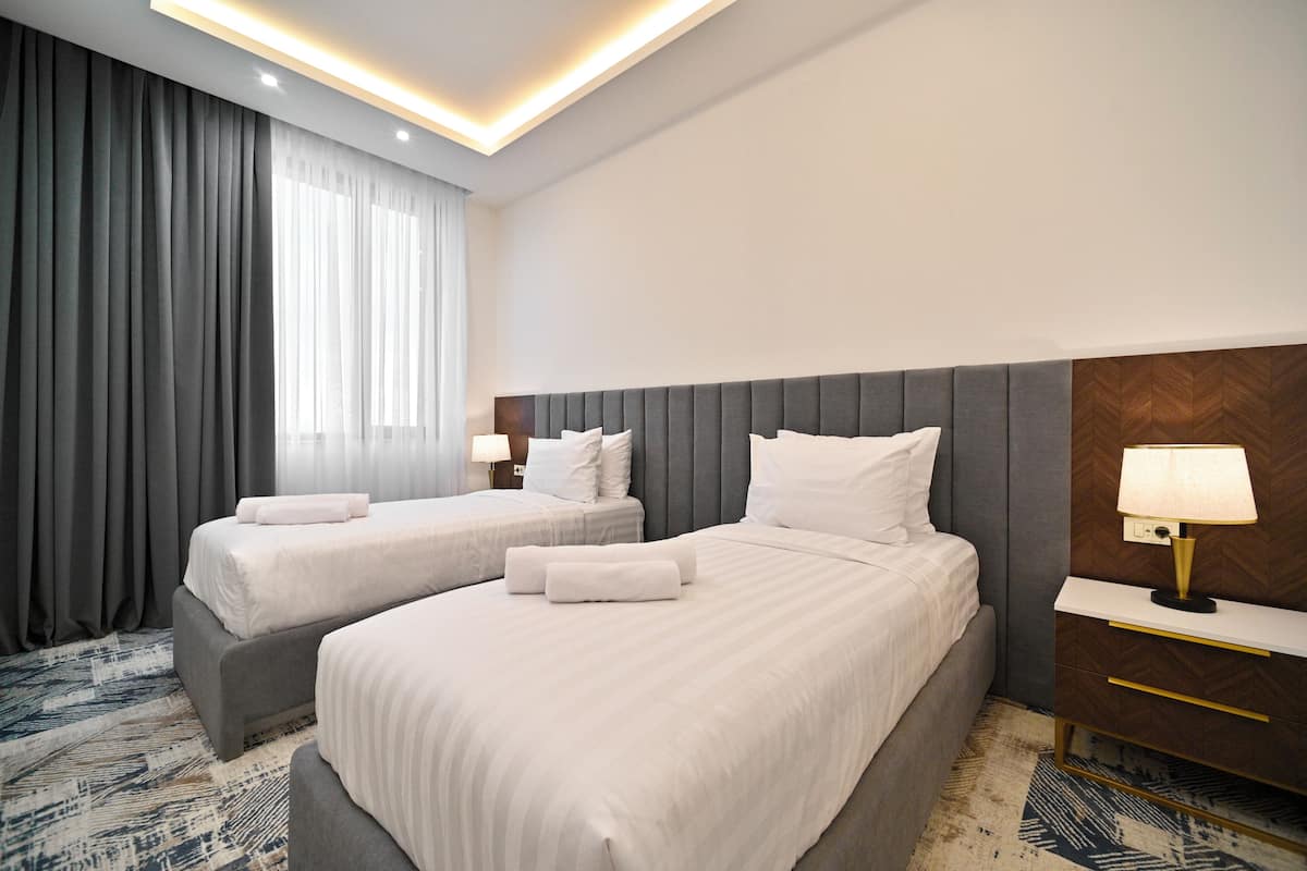 A hotel room with two beds, a nightstand, a lamp, and a large window with curtains.