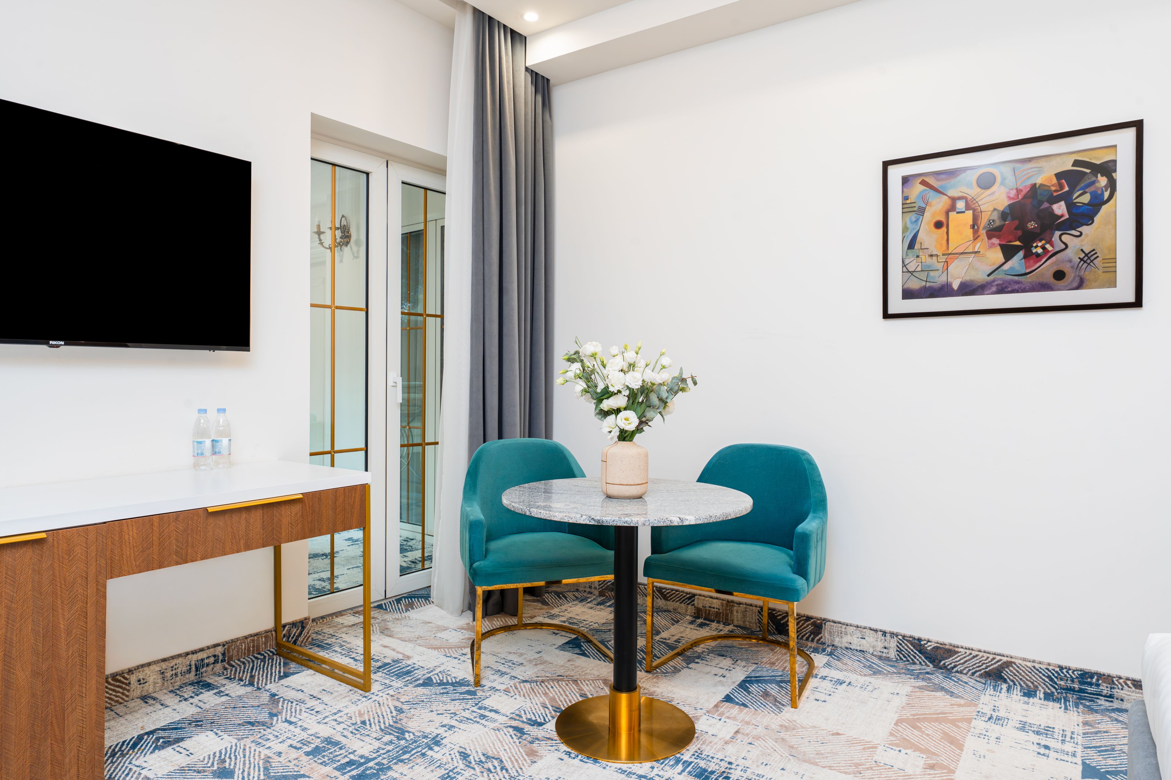 Quarto de hotel com televisão de tela plana, uma mesa redonda com cadeiras, um quadro na parede e um tapete estampado.
