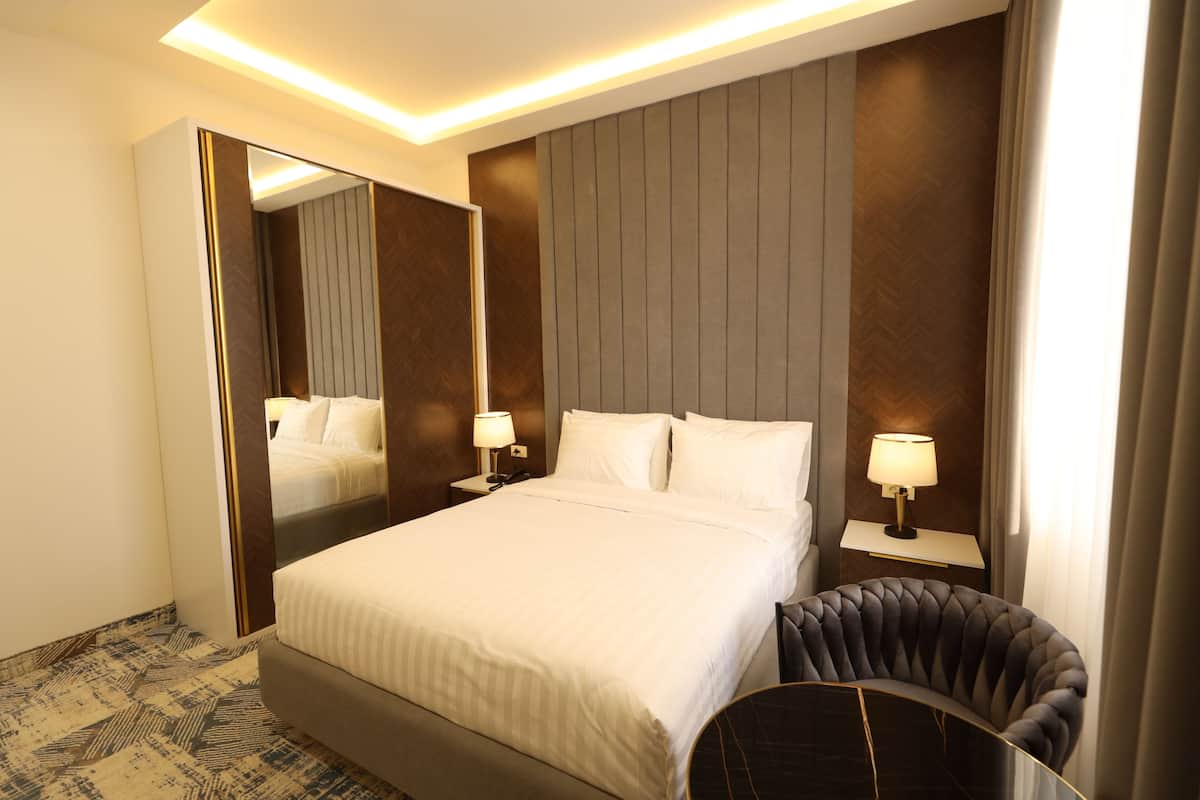 A hotel room with a large bed, two bedside tables with lamps, a mirror, and a curved armchair.