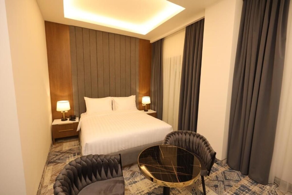 A hotel room with a bed, two bedside tables with lamps, a round coffee table, and two upholstered chairs.