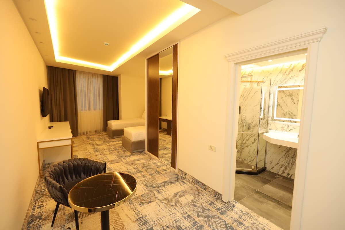 A hotel room with a bed, a small table, a chair, a desk, a mirror, and a bathroom with a marble sink.